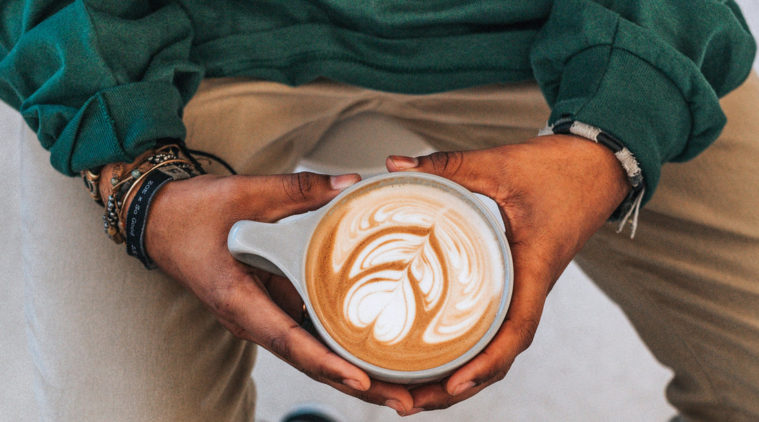 A brown skinned man holding a latte art coffee while sitting. It is paired with a Forage hyperfoods blog of "Mushroom Coffee 101: Everything you Need to Know". 