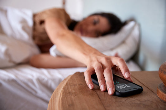 A woman lying in bed reaches out to turn off her smartphone alarm at 7:30 AM. The image captures a sleepy morning moment, emphasizing the struggle of waking up.