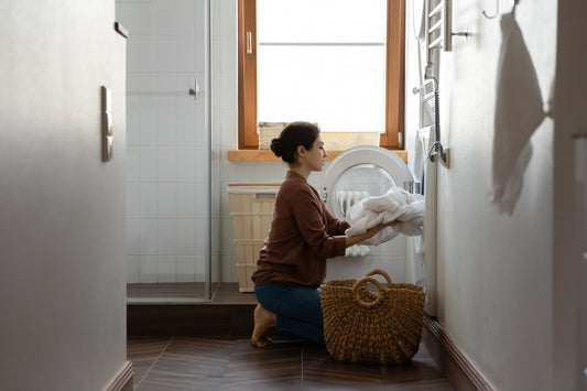 Young woman loading white laundry into front loading washing machine, doing household activities in light bathroom, productivity concept.