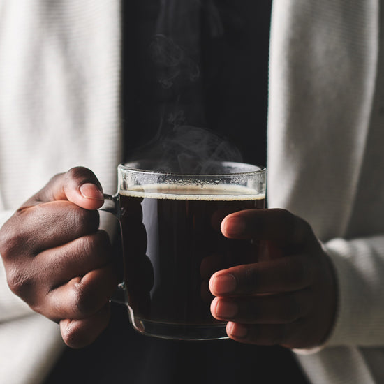 A pair of hands holding a transparent and steaming mug of dark Chaga-infused coffee. 