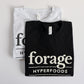 Two shirts with the word mark "Forage Hyperfoods" printed on them. The one in the background is a light gray colour with black text, the one in the foreground is black with white text.