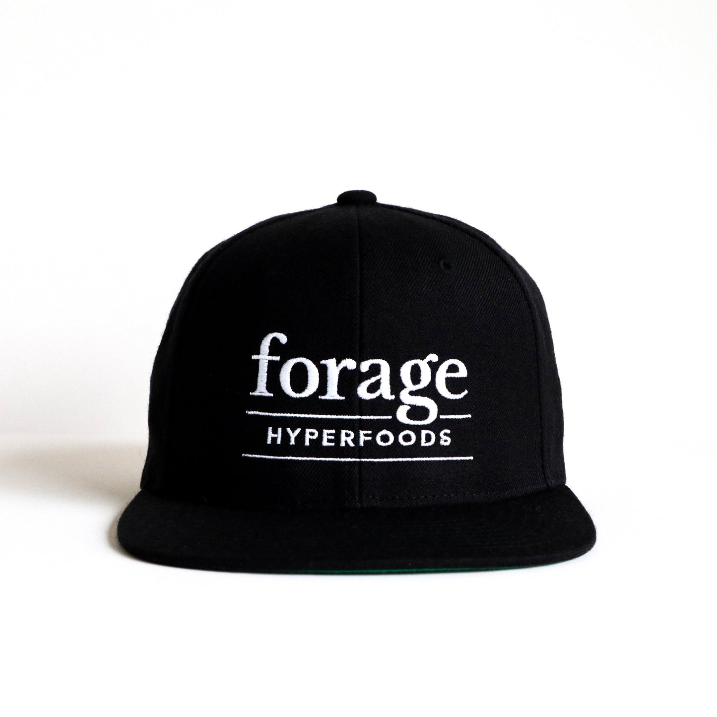 A black snapback hat with white "forage hyperfoods" embroidered on the front. It sits facing the camera. 