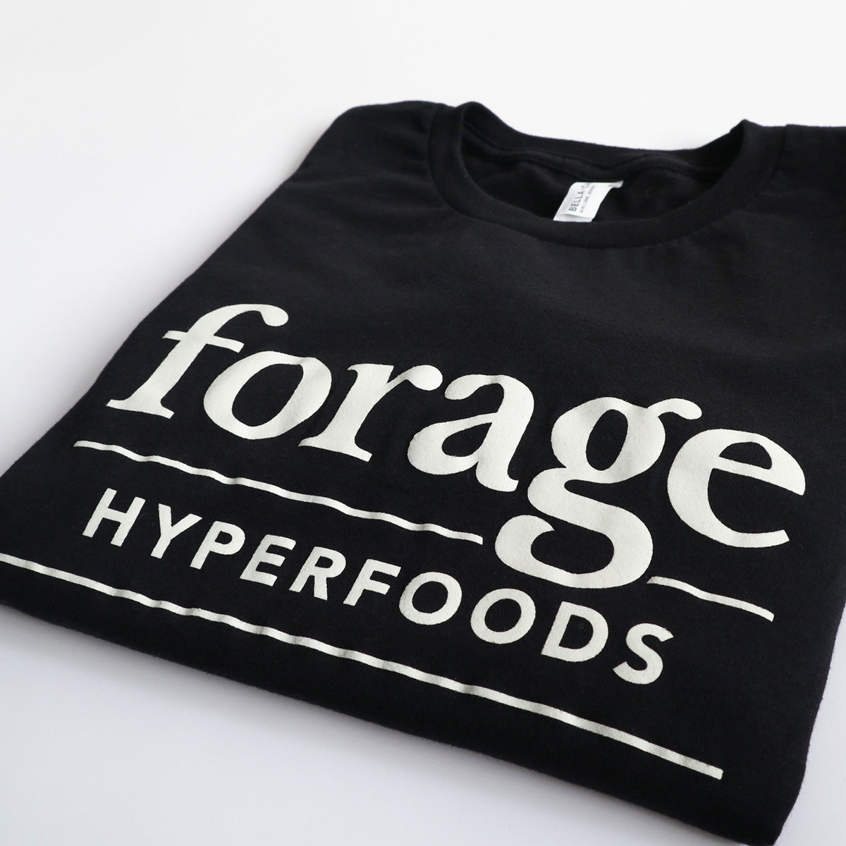 A black shirt with the word mark "Forage Hyperfoods" printed on it in white. It is laid out flat on a white background.  It is folded. 