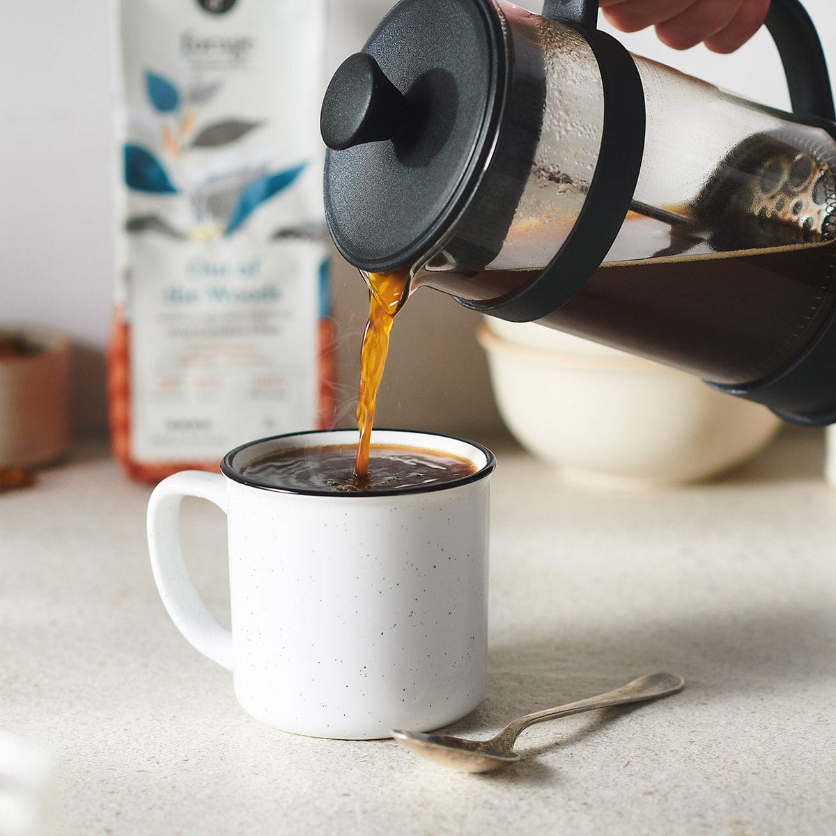 A picture of a French press pouring coffee into a plain coffee mug with the bag of Chaga-infused Out of the Woods coffee by Forage Hyperfoods in the background. 