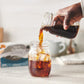 A hand pouring a brewed mason jar of chaga-infused coffee into a glass with ice cubes to make iced coffee.