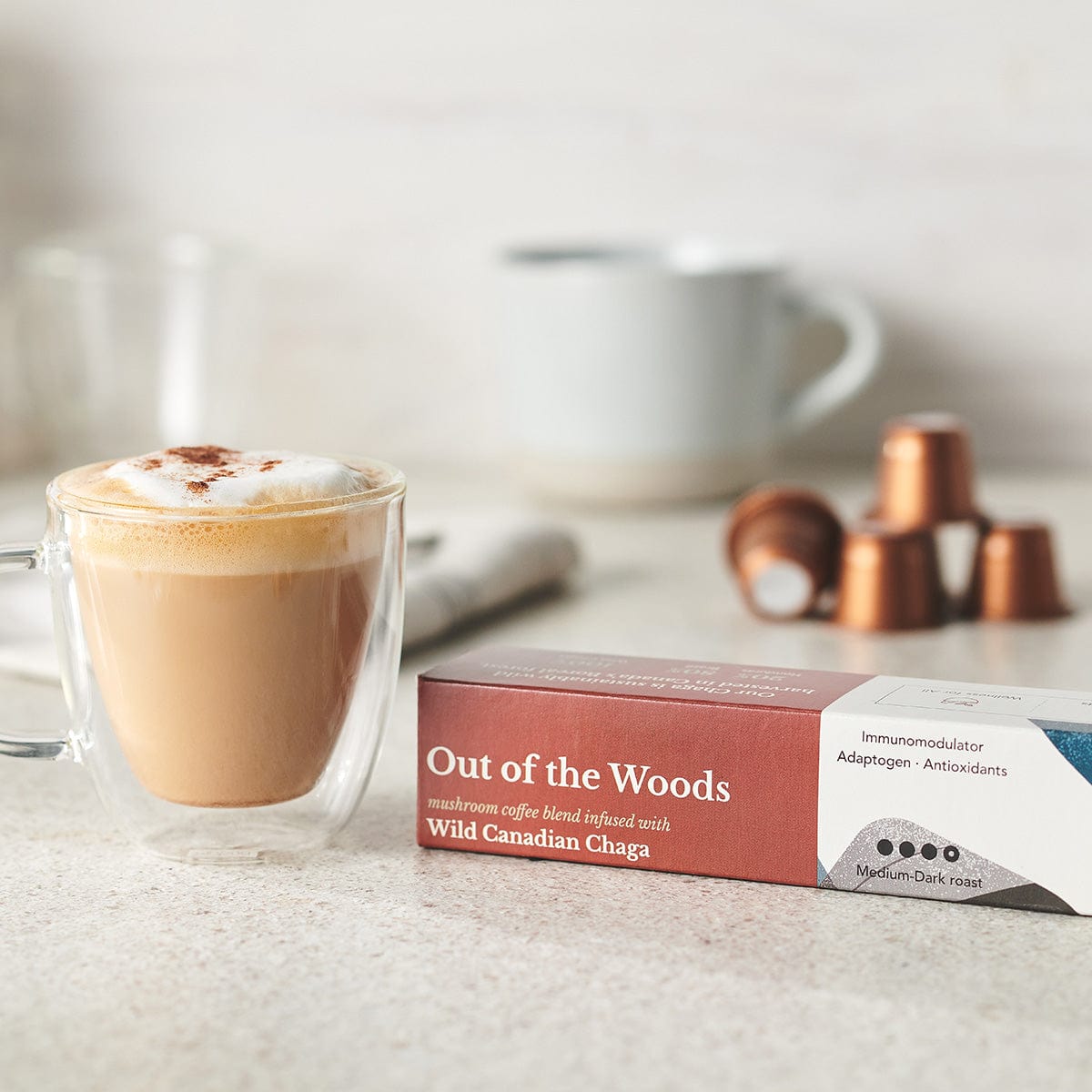 An out of the Woods Chaga-Infused Coffee in a Nespresso compatible format by Forage Hyperfoods. Next to it is a transparent mug of creamy coffee.
