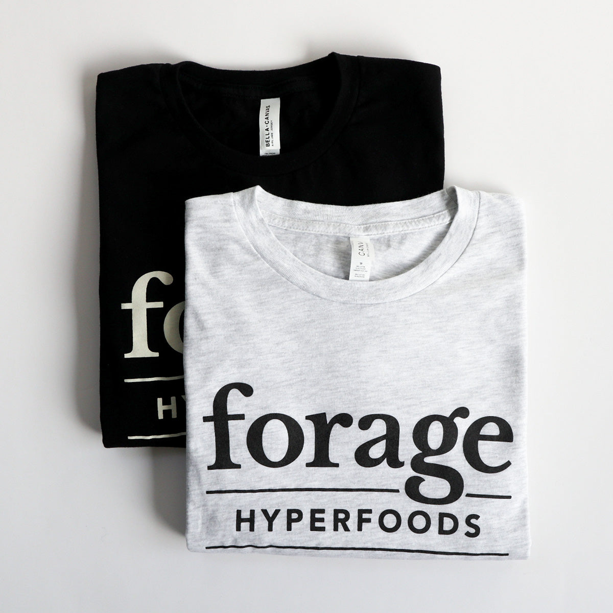 Two shirts with the word mark "Forage Hyperfoods" printed on them. The one in the foreground is a light gray colour with black text, the one in the background is black with white text.  